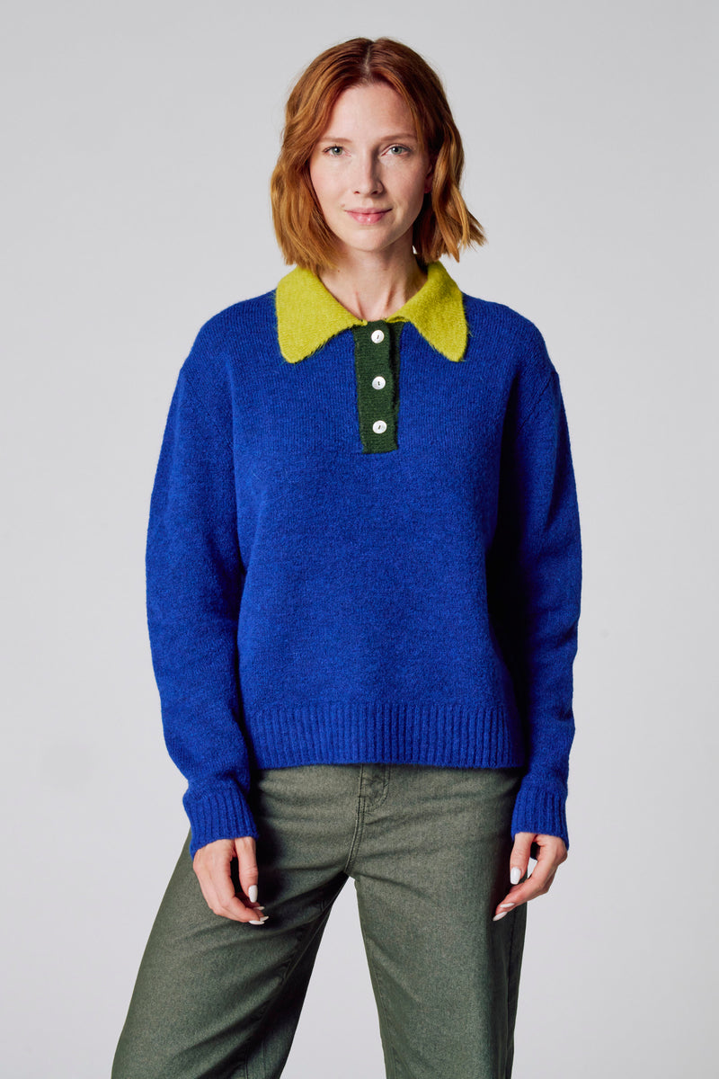 Pull à col rond bleu de COPINES D'ANVERS, avec un col vert anis et des boutons, porté avec un pantalon kaki.
