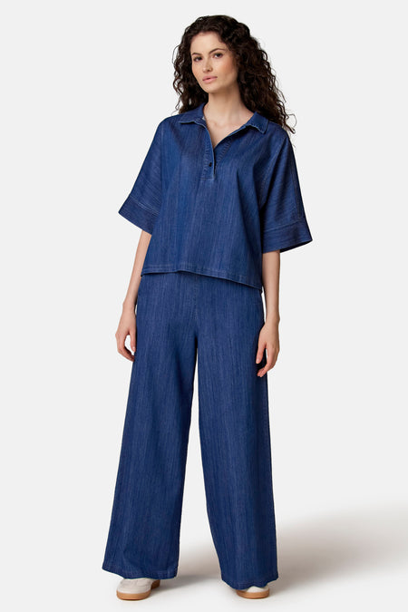 Blouse denim bleue de COPINES D'ANVERS à col polo, manches courtes et coupe décontractée, portée avec un pantalon large assorti.