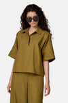 Blouse verte à manches courtes de COPINES D'ANVERS avec col à revers et décolleté fendu, coupe ample, portée avec un pantalon assorti et des lunettes de soleil.