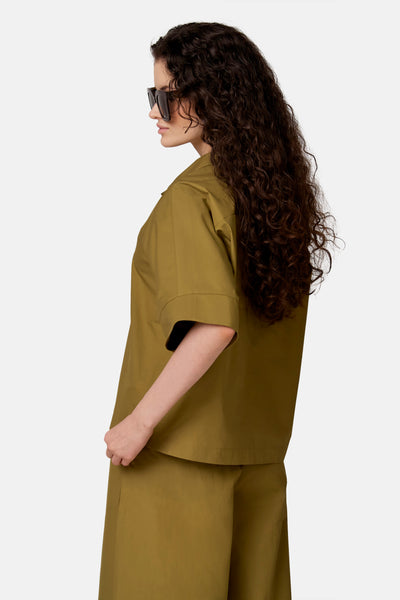 Blouse vert kaki de COPINES D'ANVERS à manches courtes et coupe ample, portée de profil avec un pantalon assorti et des lunettes de soleil.