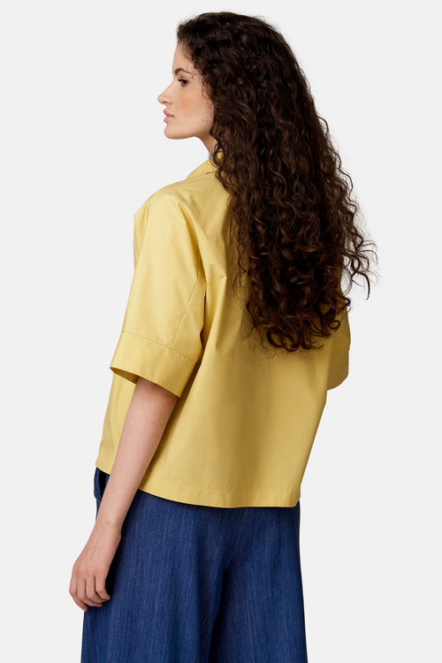 Blouse jaune à manches courtes de COPINES D'ANVERS, portée de dos par une mannequin aux cheveux bouclés, avec un pantalon ample bleu.