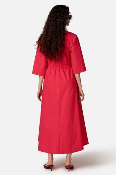 Roze lange jurk van COPINES D'ANVERS, wijdvallend, met driekwartmouwen en elastische taille, van achteren gezien met een zonnebril en muiltjes.