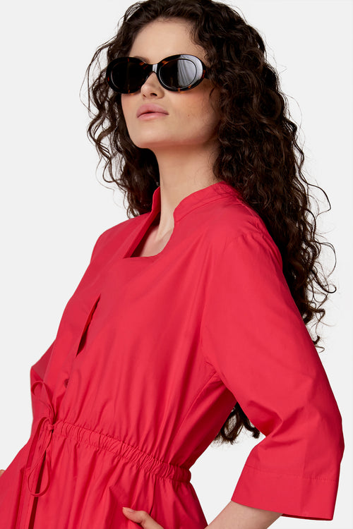 Roze jurk van COPINES D'ANVERS met opstaande kraag, vierkante halslijn, aansnoerbare taille en zakken, gedragen door een vrouw met een zonnebril.