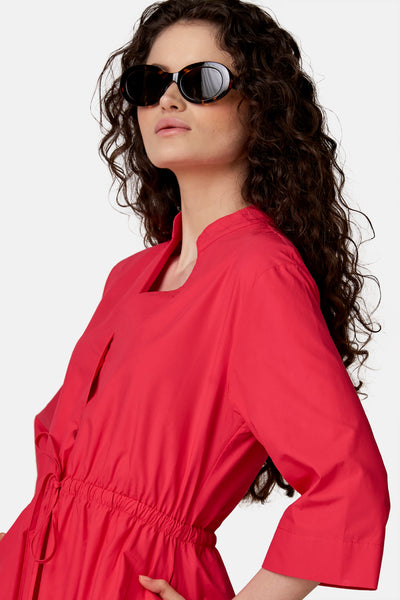 Roze jurk van COPINES D'ANVERS met opstaande kraag, vierkante halslijn, aansnoerbare taille en zakken, gedragen door een vrouw met een zonnebril.