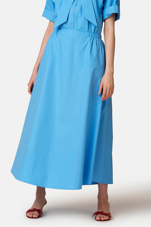 Blauwe A-lijn rok van COPINES D'ANVERS met elastische tailleband, maxi-lengte, gedragen met rode muiltjes.