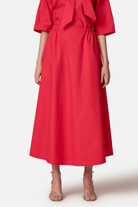 Jupe longue rouge de COPINES D'ANVERS à taille élastiquée, portée avec un haut assorti à nœud et des sandales métallisées.