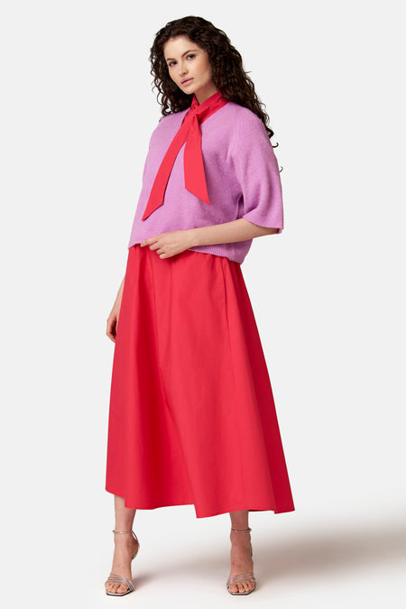 Jupe midi rouge corail de COPINES D'ANVERS portée avec un pull lavande et un nœud-lavallière rouge corail.