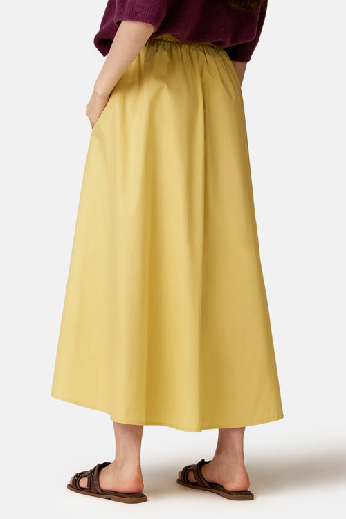 Gele midi-jurk met wijde rok van COPINES D'ANVERS met elastische taille, achterkant, gedragen met een bordeauxrode trui en bruine sandalen.