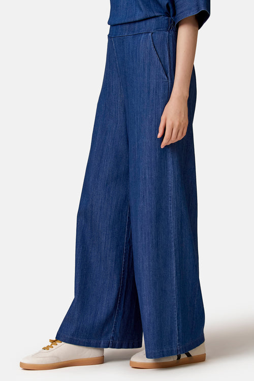Pantalon large en jean bleu de COPINES D'ANVERS, avec taille élastiquée et poches latérales, porté avec des baskets claires.