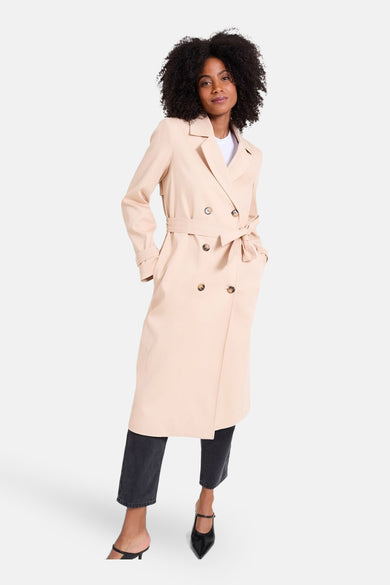 Veste longue beige par NAF NAF, à double boutonnage avec ceinture à nouer, portée avec un jean noir.

