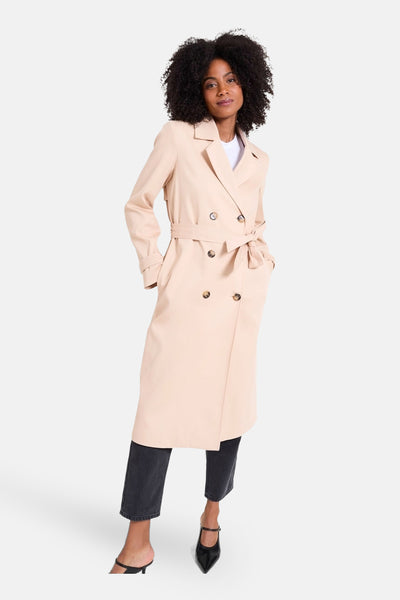 Veste longue beige par NAF NAF, à double boutonnage avec ceinture à nouer, portée avec un jean noir.
