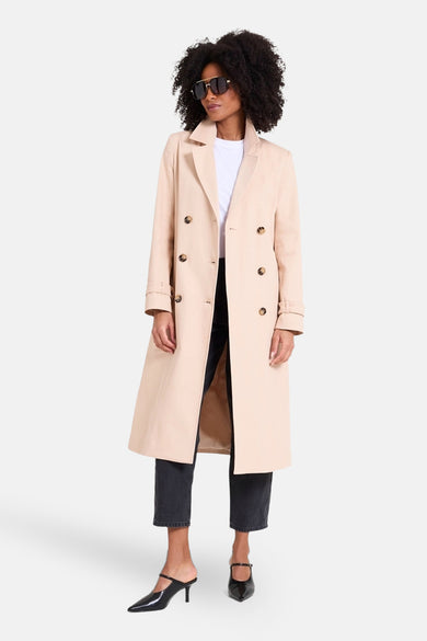 Veste longue beige de NAF NAF, à double boutonnage, portée avec un jean noir et des chaussures à talons noires.