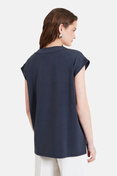 T-shirt gris à manches courtes de NAF NAF, coupe ample, porté avec un pantalon blanc et des boucles d'oreilles argentées.