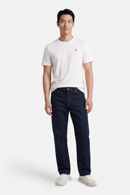 Ecru T-shirt van STROM met een klein rood logo op de borst, gedragen door een man met een donkere spijkerbroek en witte sneakers.