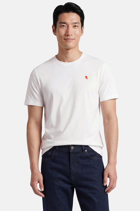 Ecru T-shirt van STROM met een klein geborduurd rood-wit stoellogo, gedragen door een man in donkerblauwe jeans.