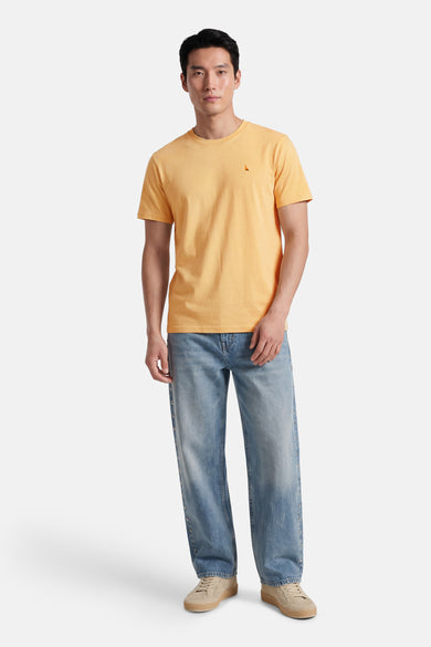 T-shirt jaune de STROM avec logo triangulaire, porté avec un jean clair à œillets et des baskets beiges perforées.