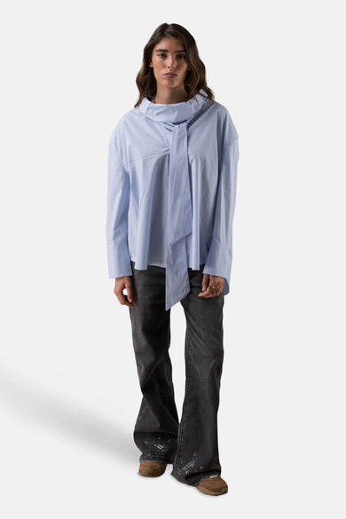 Chemise à manches longues bleue de ADORE, avec un col à nouer et portée avec un jean foncé et des chaussures marron.