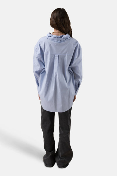 Chemise à manches longues bleue par ADORE, avec un collier doré, portée avec un pantalon noir.
