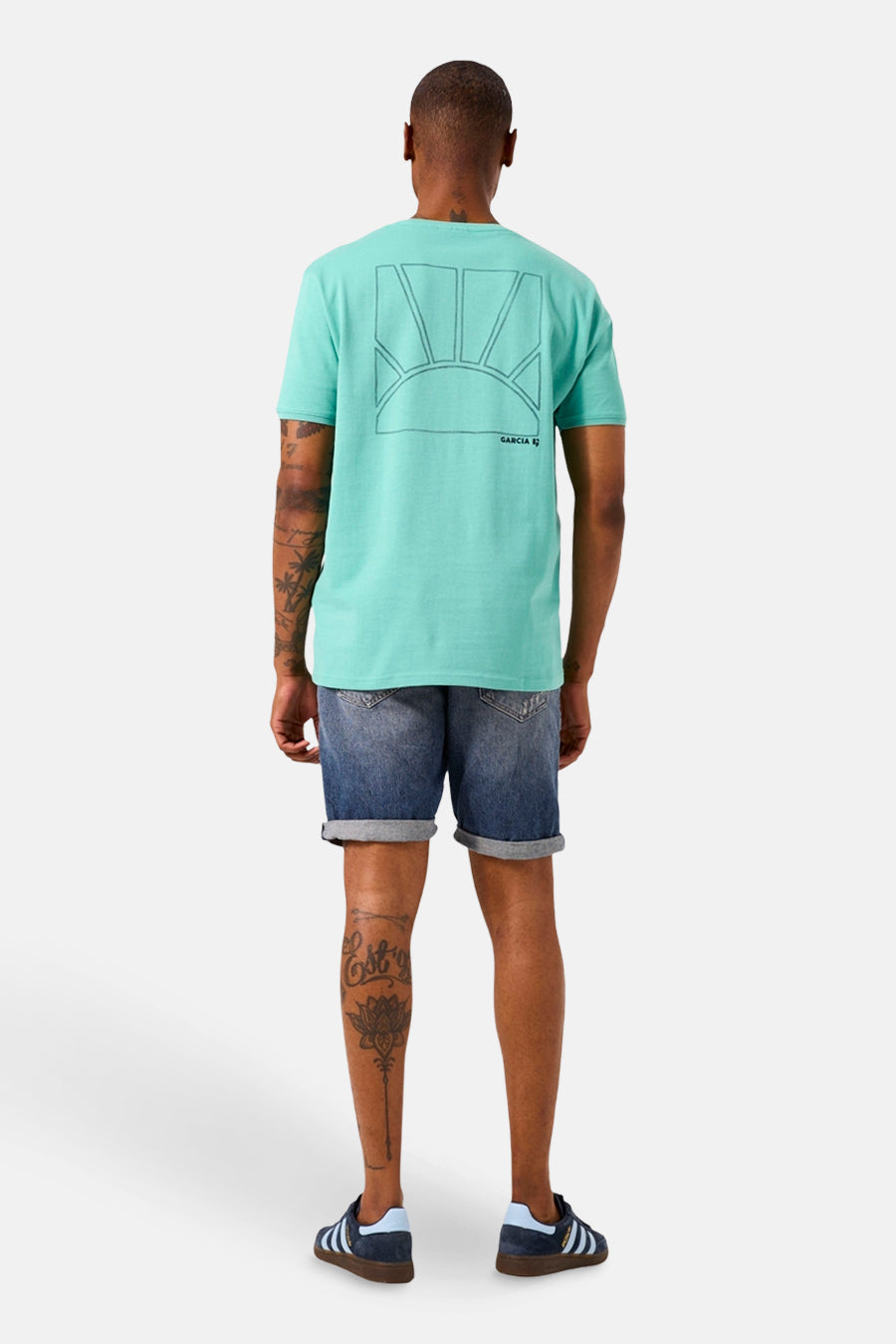 T-shirt à manches courtes vert de GARCIA, imprimé soleil au dos, porté avec un short en jean délavé et des baskets bleues.
