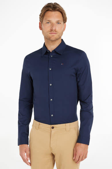 Chemise à manches longues bleue de Tommy Jeans, avec des boutons et portée avec un pantalon beige.
