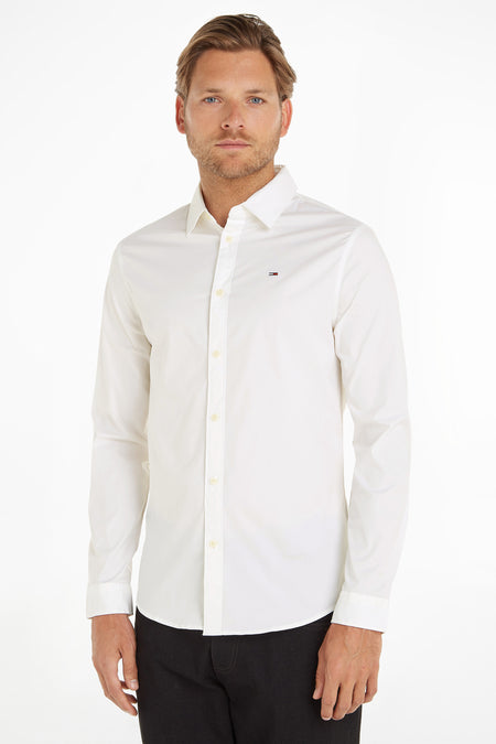 Chemise blanche à manches longues de Tommy Jeans, portée par un homme, avec un pantalon noir visible.