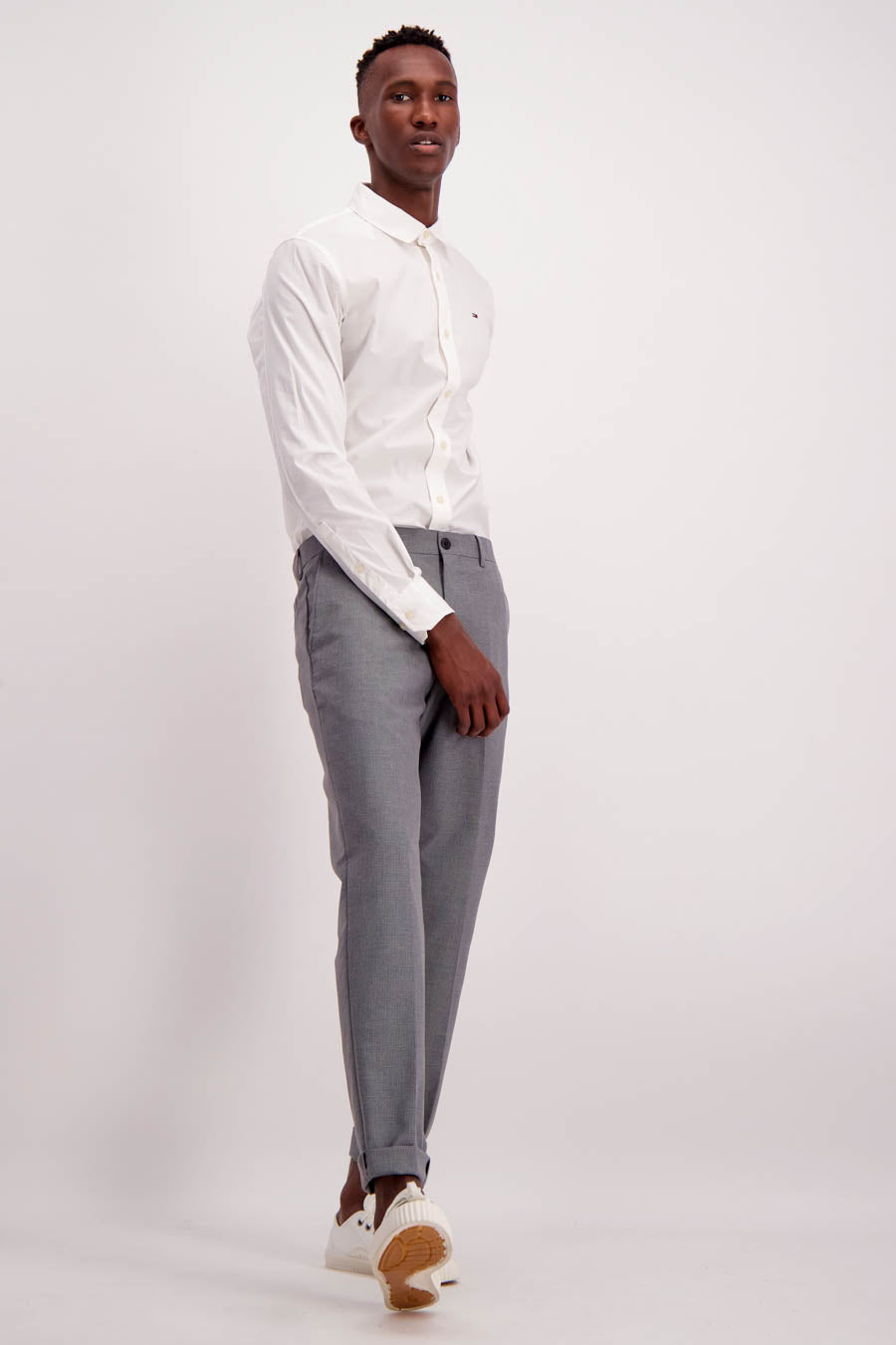 Chemise blanche à manches longues de Tommy Jeans, avec une coupe classique et un col boutonné, portée avec un pantalon gris.
