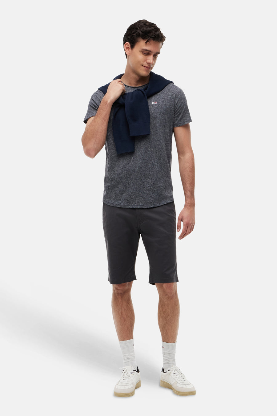 T-shirt gris chiné à manches courtes de Tommy Jeans, avec un petit logo sur la poitrine. Un pull bleu marine est drapé sur les épaules du modèle.