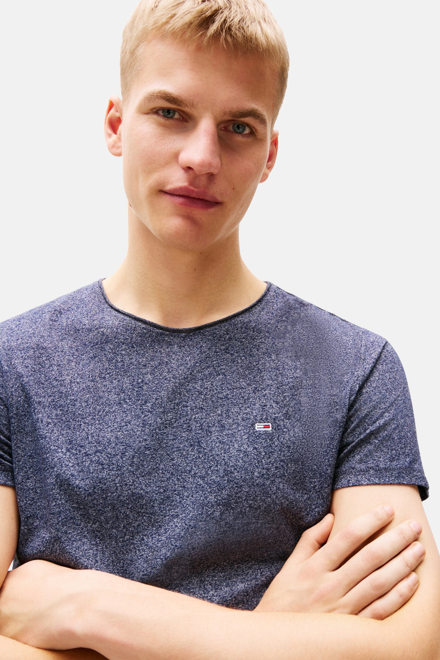 T-shirt bleu chiné à manches courtes de Tommy Jeans, porté par un homme aux bras croisés.