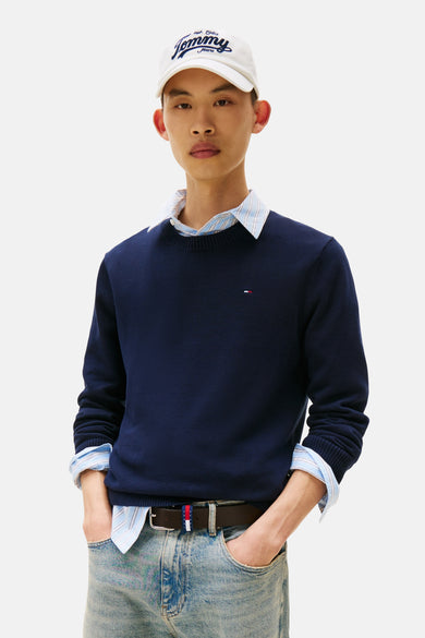 Pull à col rond bleu foncé de Tommy Jeans, porté sur une chemise rayée et avec un jean clair.
