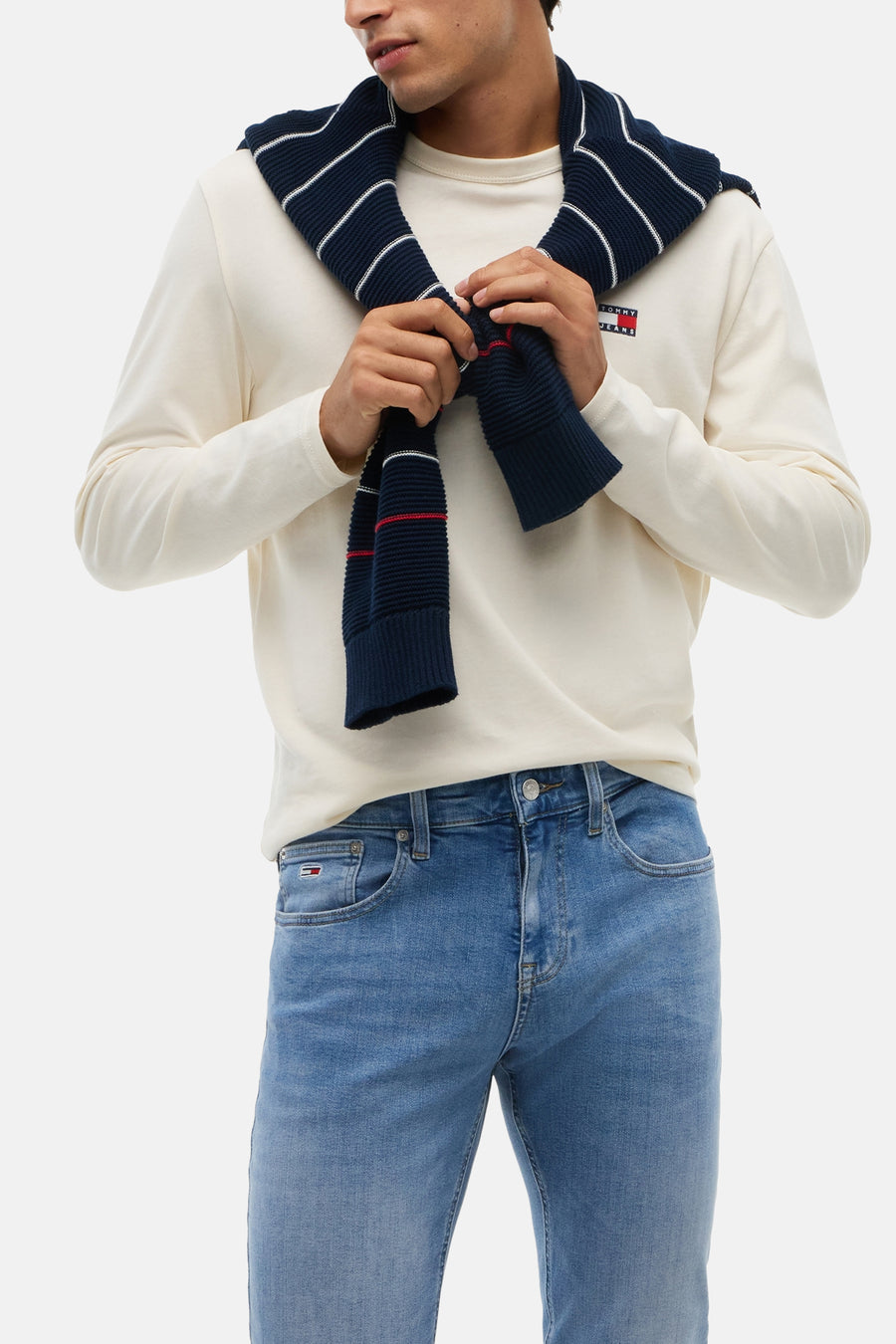 Light blue slim-fit denim jeans by TOMMY JEANS, featuring faded wash and front pocket branding, worn with cream long-sleeve tee and navy striped scarf.