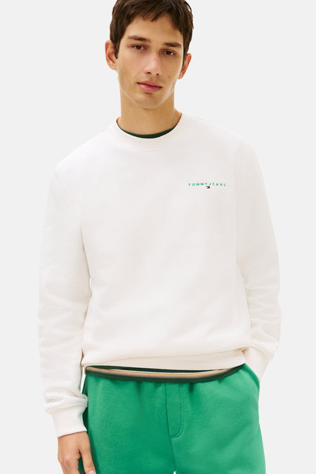 Sweat-shirt blanc col rond de TOMMY JEANS, avec logo discret sur la poitrine, porté avec un pantalon de survêtement vert.
