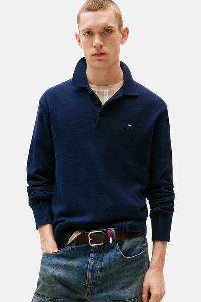 Pull bleu par Tommy Jeans avec un col polo, porté sur une chemise rayée beige et un jean bleu.
