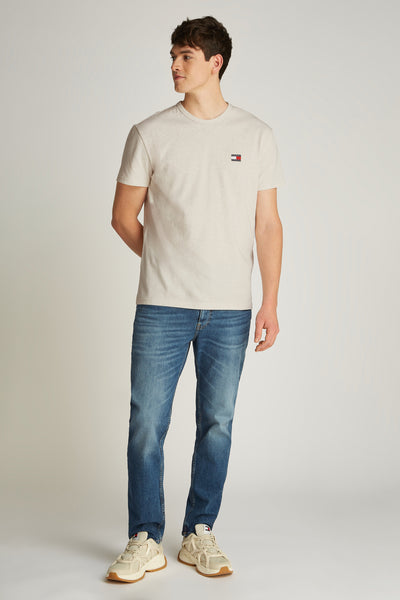 Ecru T-shirt met korte mouwen van TOMMY JEANS, met logo op de borst, gedragen met een blauwe jeans en beige sneakers.