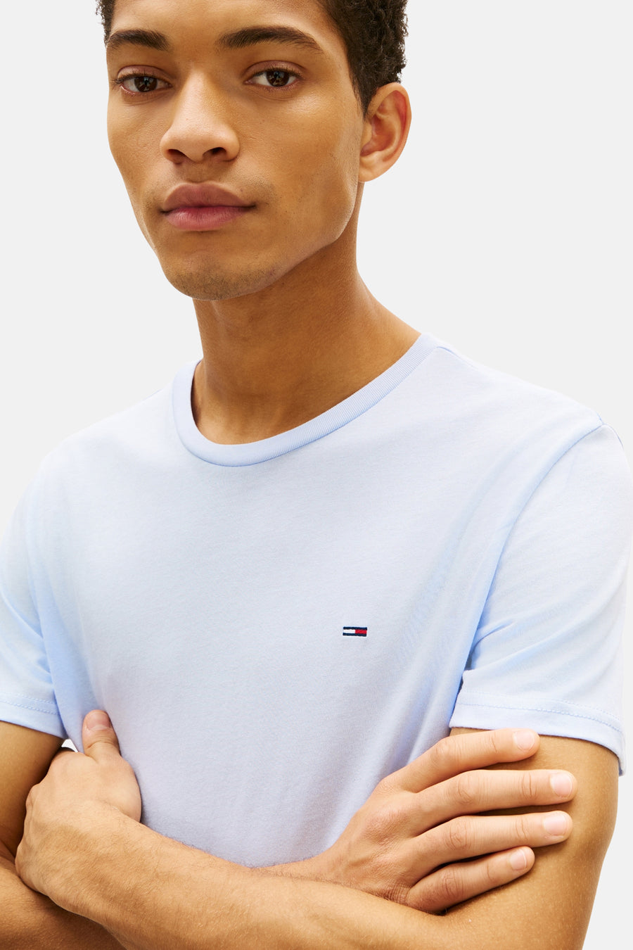 T-shirt bleu à manches courtes de TOMMY JEANS, avec logo discret sur le devant, porté par un homme, les bras croisés.