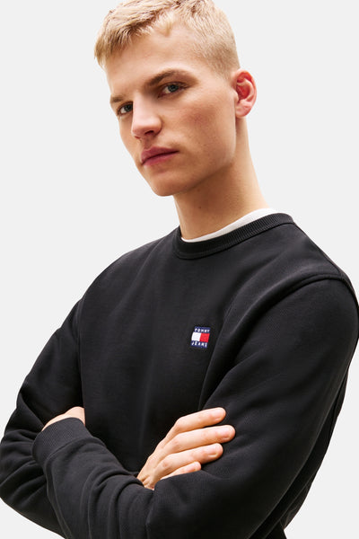 Sweat noir col rond de TOMMY JEANS, porté avec un t-shirt blanc à col rond visible au cou.