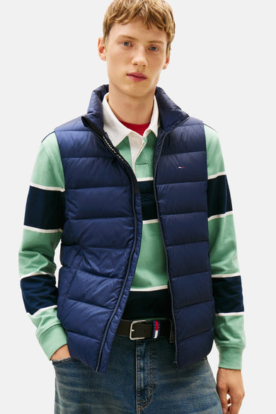 Blauwe mouwloze jas van TOMMY JEANS, gedragen over een gestreept poloshirt en een wijde jeans met riem.