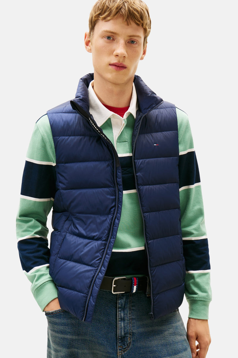 Blauwe mouwloze jas van TOMMY JEANS, gedragen over een gestreept poloshirt en een wijde jeans met riem.