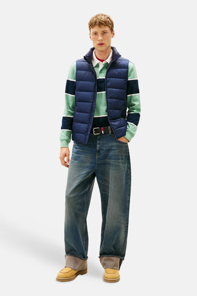 Veste sans manches bleue de TOMMY JEANS, portée sur un polo rayé et un jean ample avec ceinture.