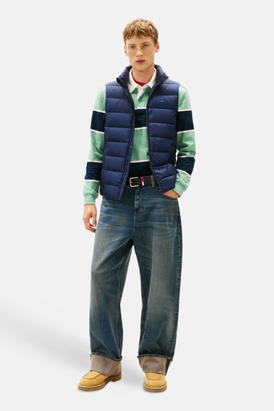 Veste sans manches bleue de TOMMY JEANS, portée sur un polo rayé et un jean ample avec ceinture.