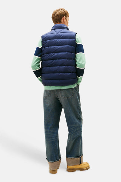 Marineblauwe mouwloze jas van TOMMY JEANS, gedragen over een gestreept T-shirt, met een spijkerbroek en mosterdkleurige laarzen.