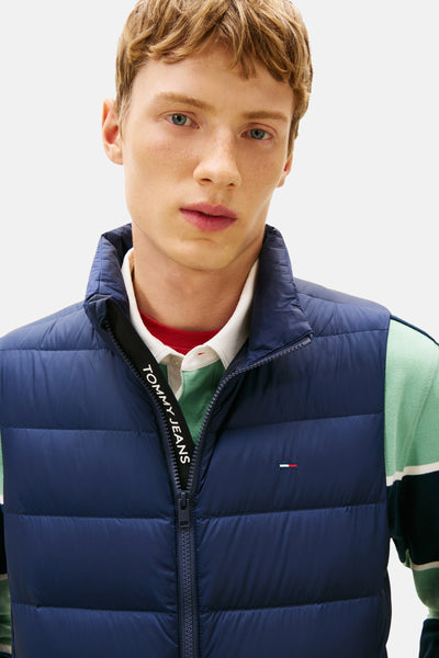 Blauwe mouwloze jas van TOMMY JEANS met zichtbare ritssluiting, gedragen over een gestreept poloshirt.