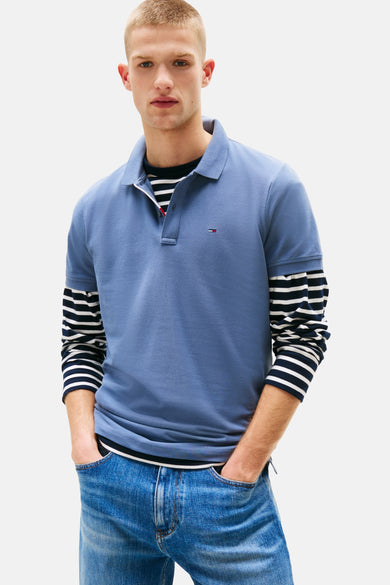 Polo à manches courtes bleu de TOMMY JEANS, porté sur un t-shirt rayé à manches longues et un jean.