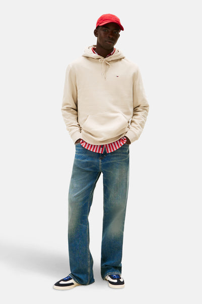 Sweat à capuche beige de TOMMY JEANS, porté sur une chemise rayée rouge et blanche, avec un jean bleu ample et une casquette rouge.
