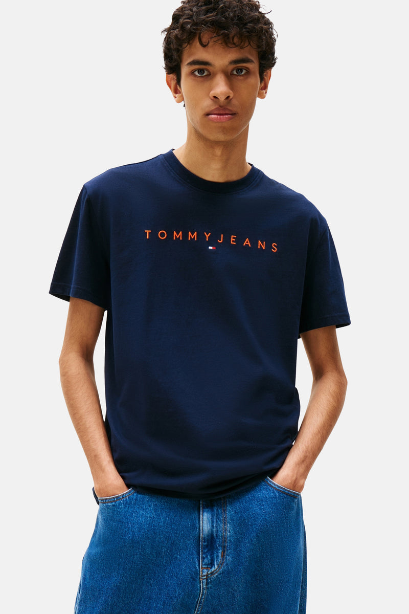 Blauw T-shirt met korte mouwen van TOMMY JEANS, met oranje opdruk aan de voorkant, gedragen met een spijkerbroek.