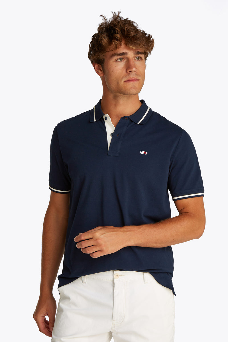 Blauw poloshirt met korte mouwen van TOMMY JEANS met witte bies op de kraag en mouwen, gedragen met een witte broek.