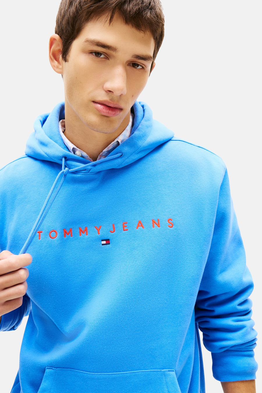 Blauwe hoodie van TOMMY JEANS met rode opdruk aan de voorkant, gedragen met een geruit overhemd.