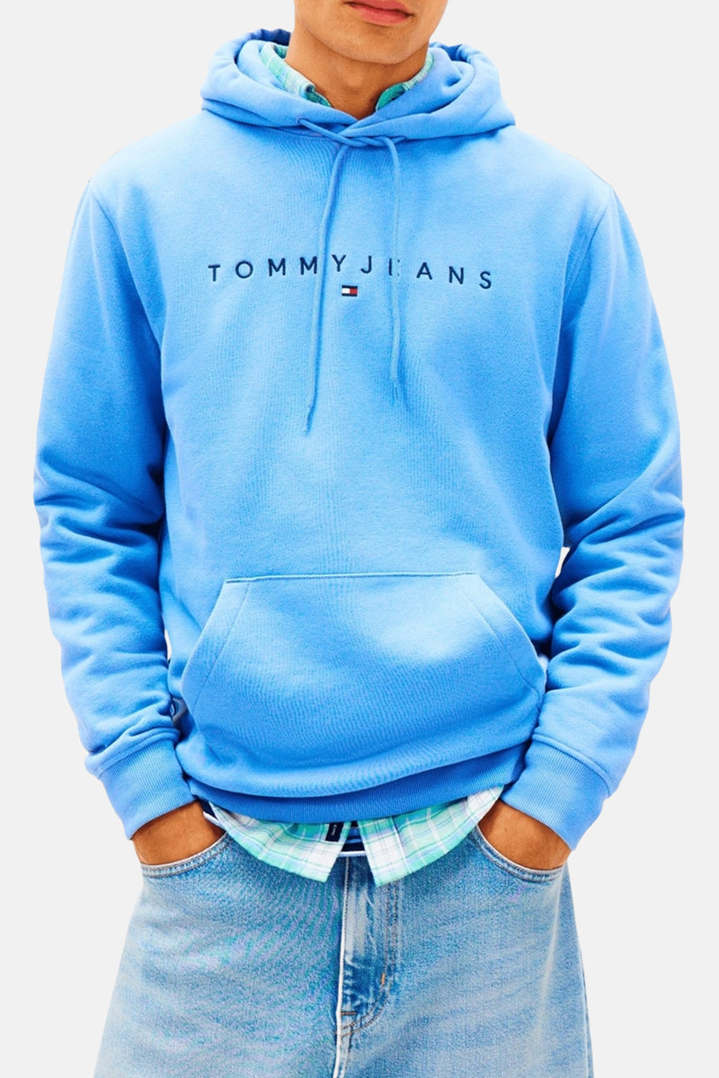 Sweat à capuche bleu clair de TOMMY JEANS avec inscription et drapeau brodés sur la poitrine, poche kangourou, et porté avec une chemise à carreaux.