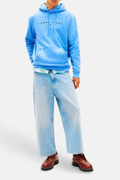 Sweat à capuche bleu de TOMMY JEANS avec logo sur la poitrine, poche kangourou, porté avec un jean large et une chemise à carreaux.