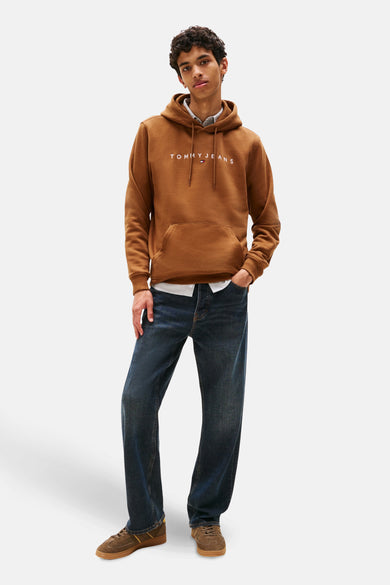 Bruine hoodie van TOMMY JEANS, met trekkoord en kangoeroezak, gedragen over een overhemd en een casual spijkerbroek.