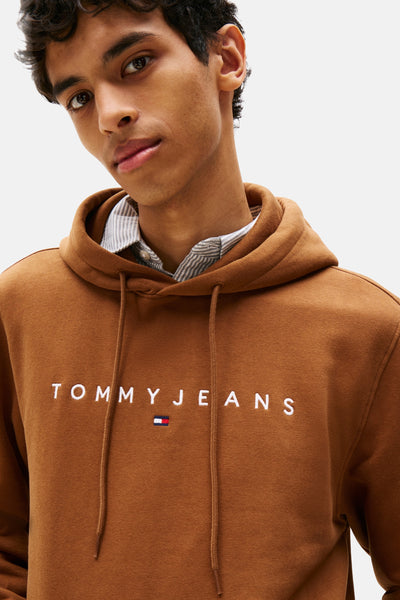 Sweat à capuche brun de TOMMY JEANS, avec cordon de serrage et logo brodé sur le devant, porté sur une chemise rayée.